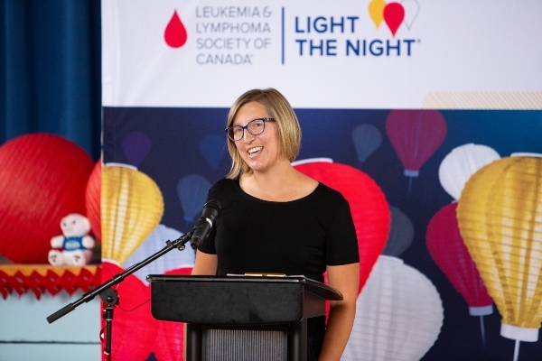 一个有深色金发鲍勃发型，眼镜和黑色上衣的女人，讲台。在她身后是加拿大白血病和淋巴瘤协会，夜晚，红色，黄色和白纸灯笼。
