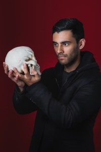 一名身穿黑色夹克的男子站在红色背景前，手里拿着一个头骨，看着它的眼窝。