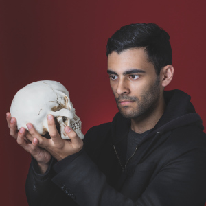 一个黑发的男人和轻微的胡子,戴着一个dark hoodie, looks intently at the human skull he holds in his hands