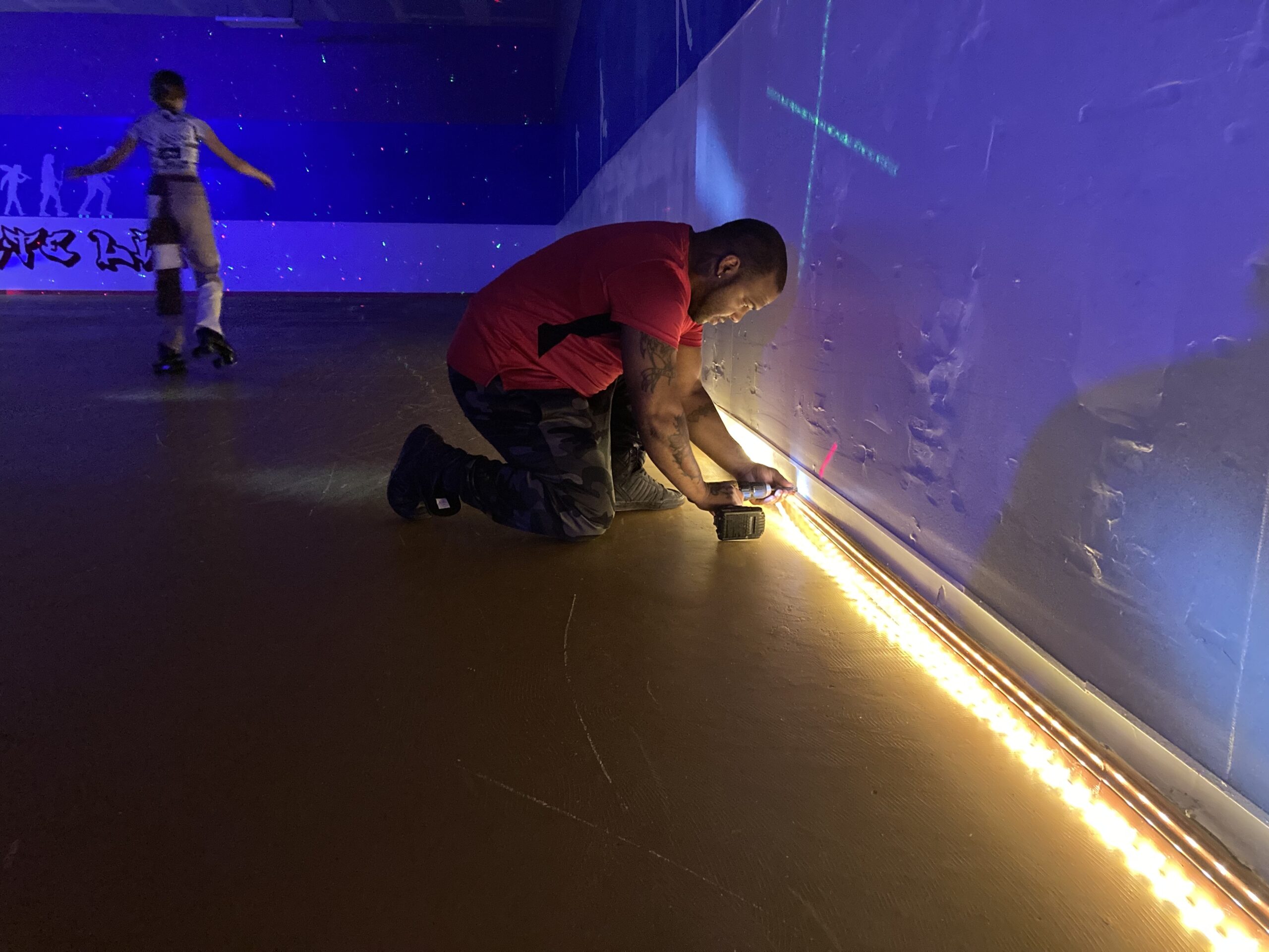 黑人穿红色T恤钻头，并在昏暗的滚筒溜冰场上安装踢脚板灯