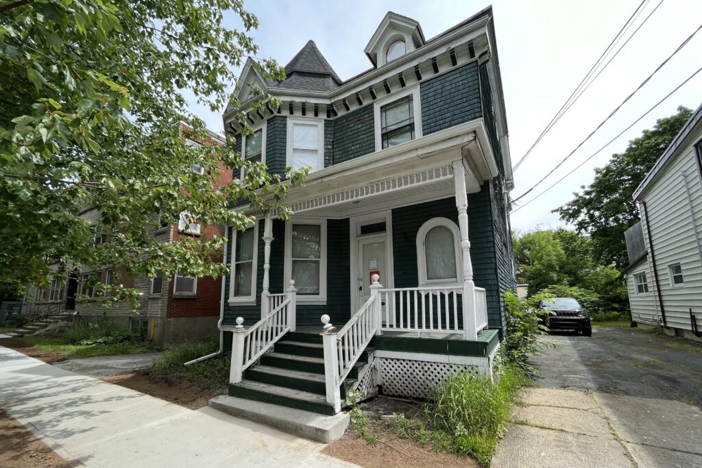 An old house with green cedar shingles and ornate white trim is seen on a sunny day.