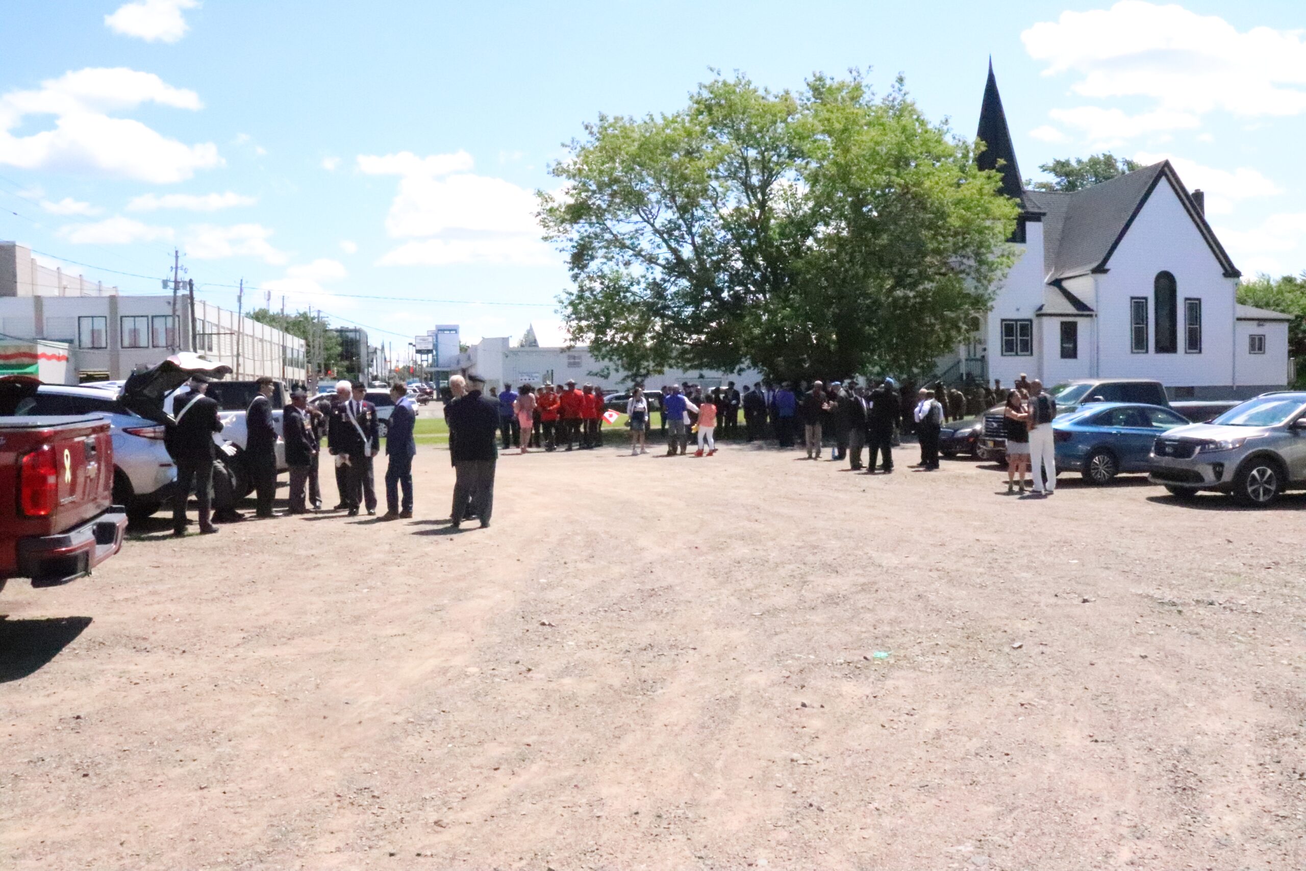 People stand in a dusty parking lot in front of a white church. A large tree stands in front of the church door.