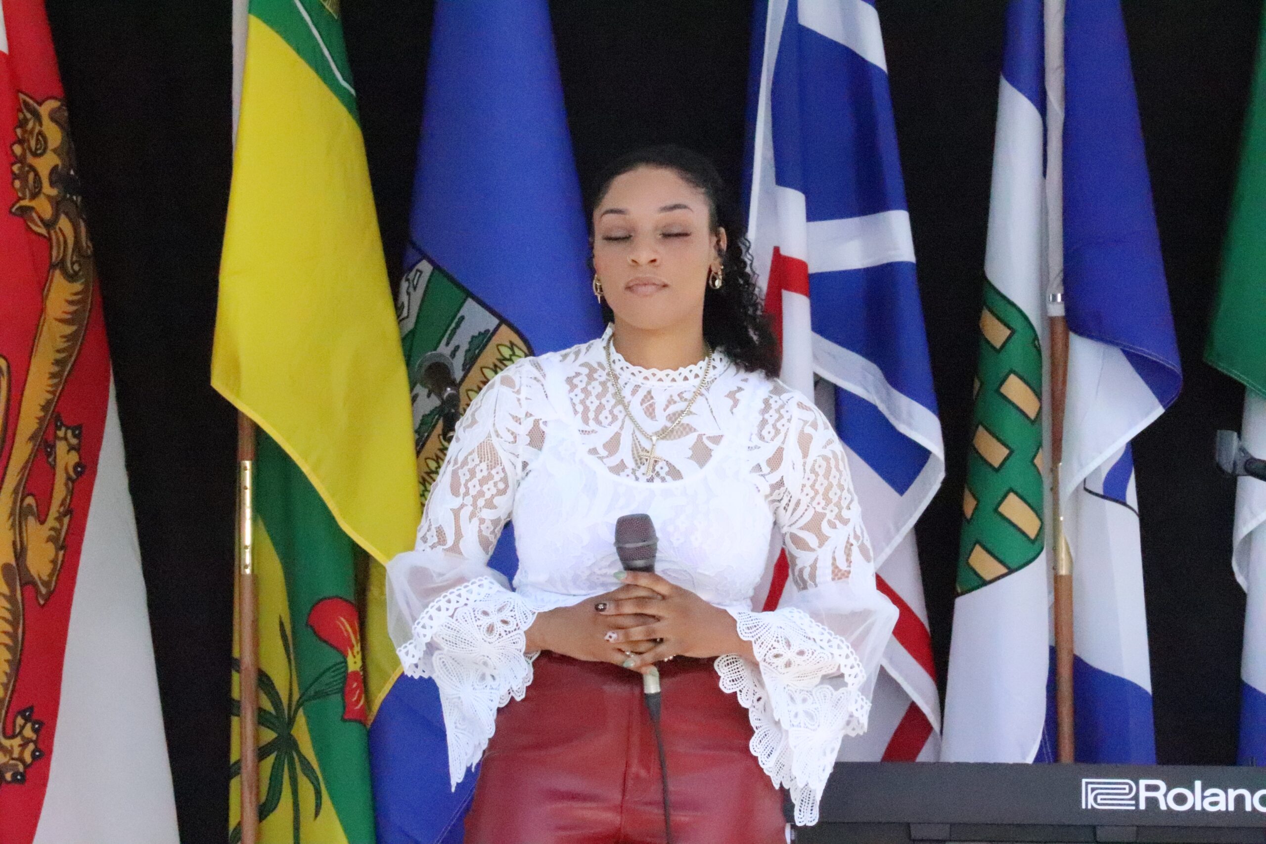 A young Black woman with a white lacey top and red pants stands with a microphone on a stage. Behind her are several flags.