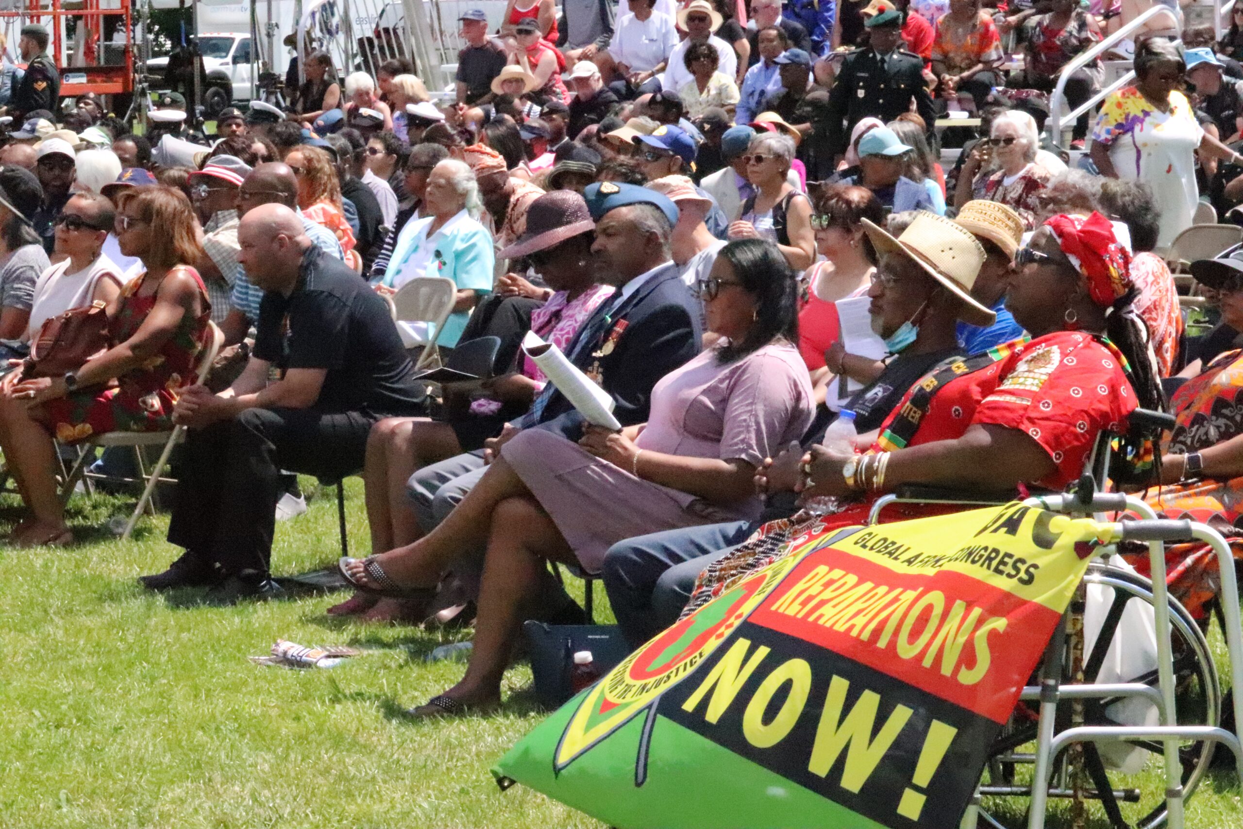 Crowds of people sit in chairs in a park watching a ceremony on a sunny day.