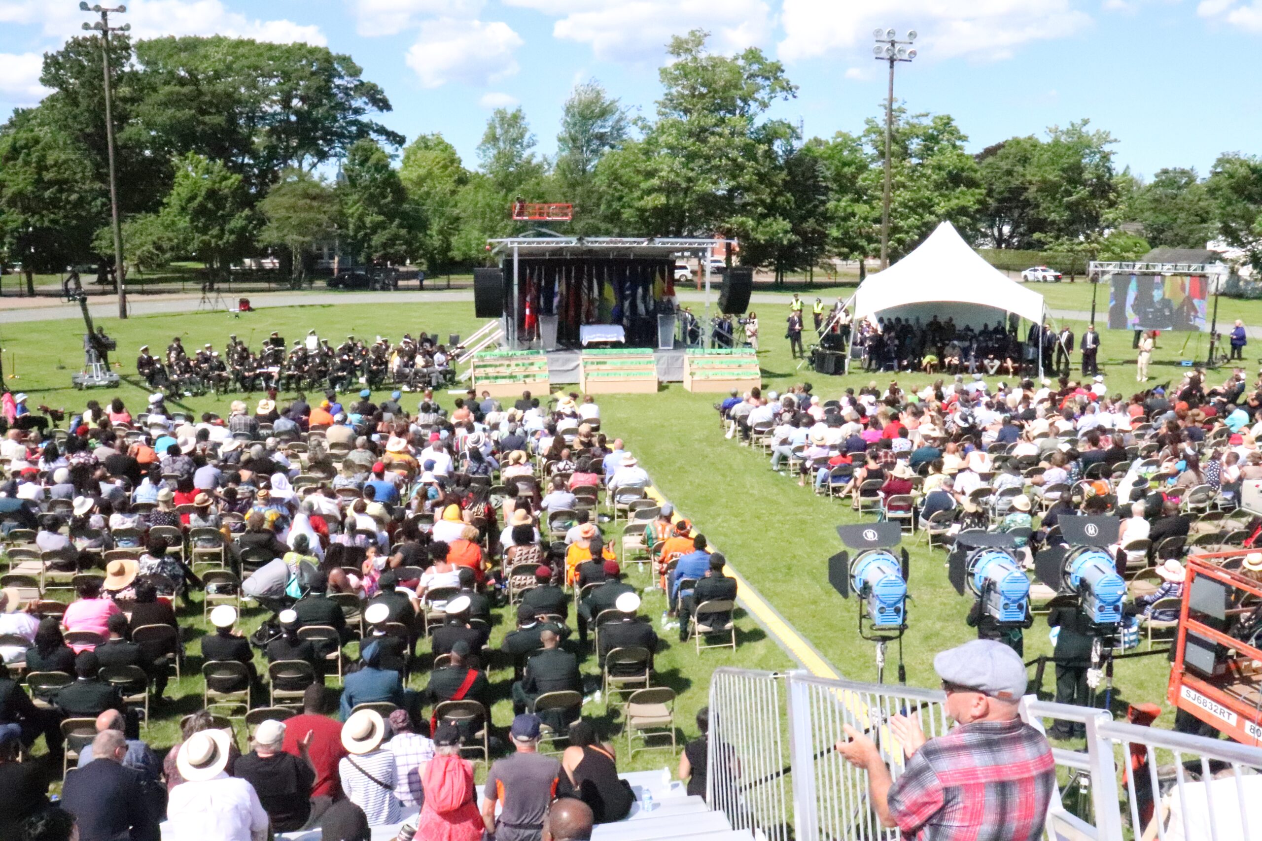 An overheard view of a crowd in a park listening to speaks on a stage.
