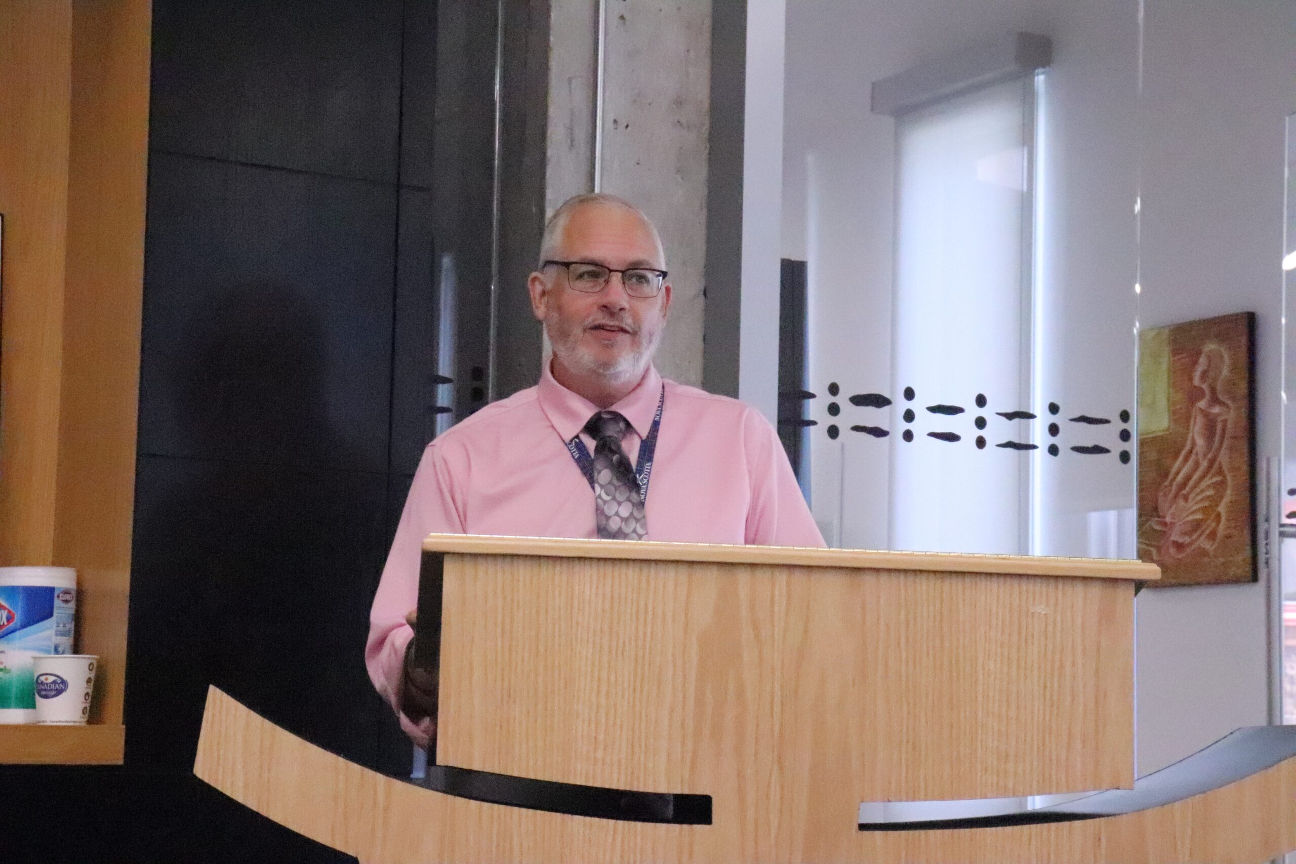 一个有灰色头发和山羊胡子的男人，穿着粉红色的衬衫，带有打印领带架在讲台上