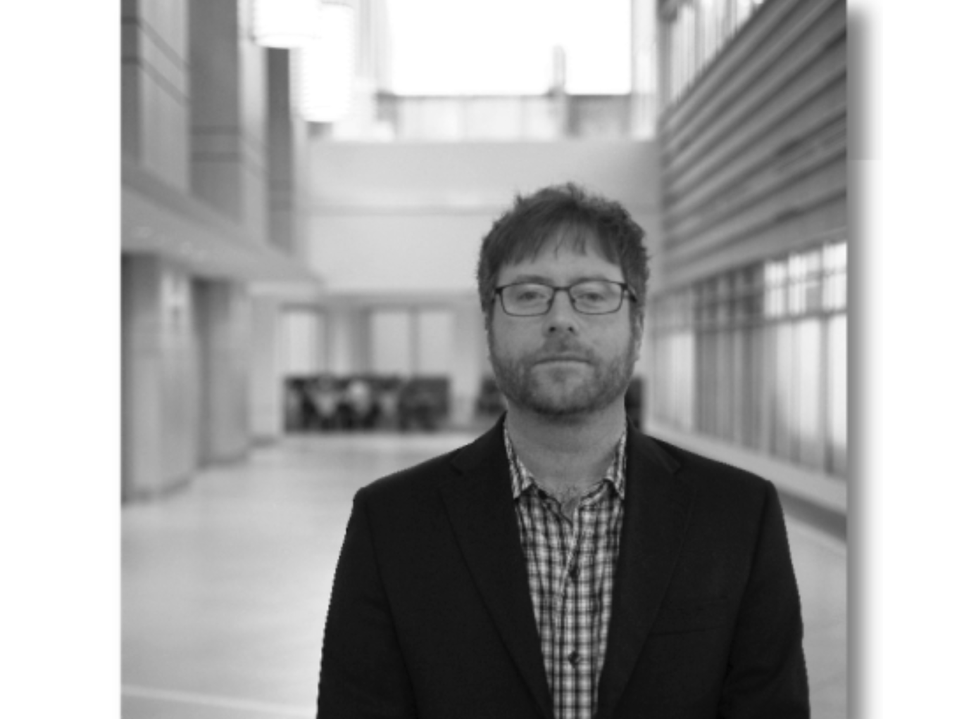 一张黑白照片上，一个戴眼镜、穿着格子衬衫和深色夹克的大胡子男子站在一个宽敞的机构门厅里。