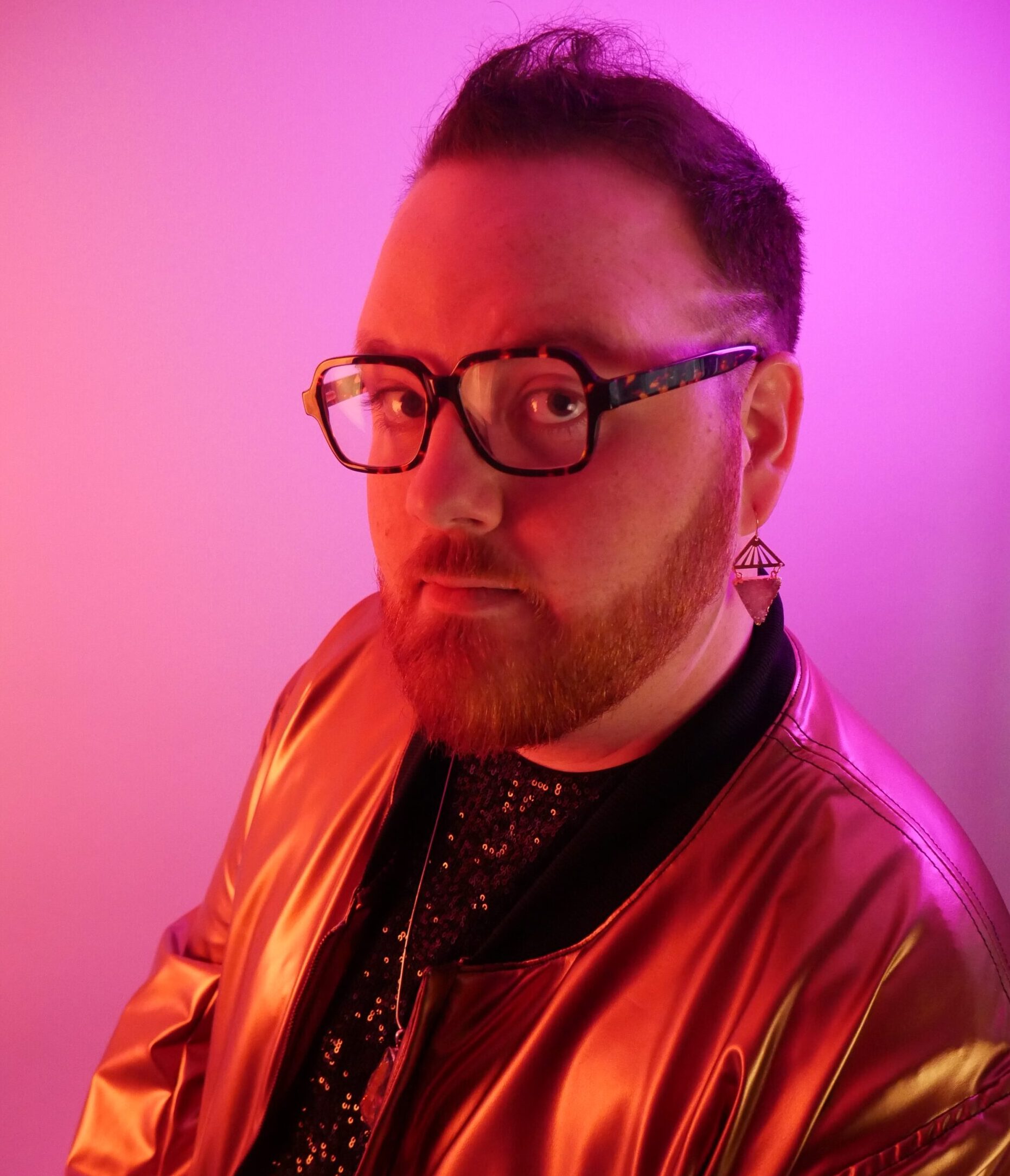 一个戴眼镜的白人男子的肖像，整齐修剪的胡子和短发，穿着薄夹克，靠向相机。