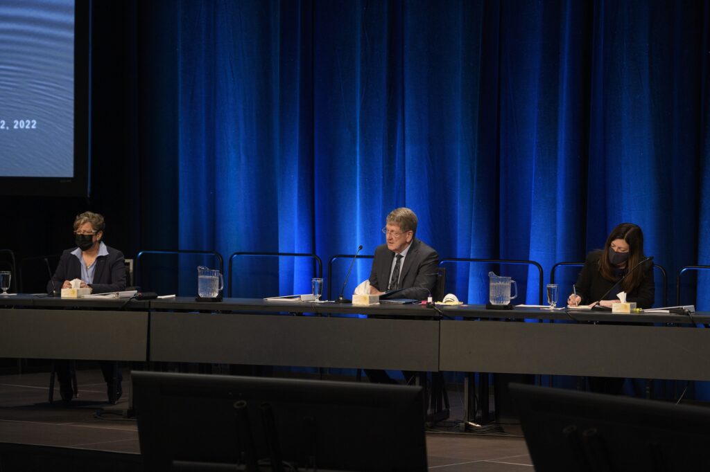 三个人坐在蓝色窗帘前的一张长桌旁。一个视频屏幕在他们的右边