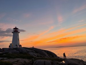 日落时分，人们走在灯塔附近的岩石上。天空被蓝色和橙色照亮了。