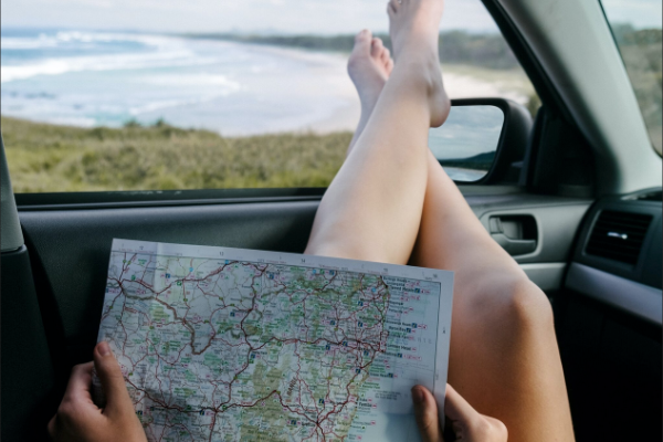 一名白人妇女双手拿着地图，在靠近海滩的车里，她的脚伸出驾驶座一侧的窗户