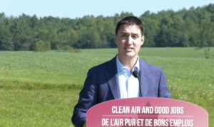 一个新剪了短发、穿着蓝色格子西装的白人男子站在以树为背景的田野里的讲台上。