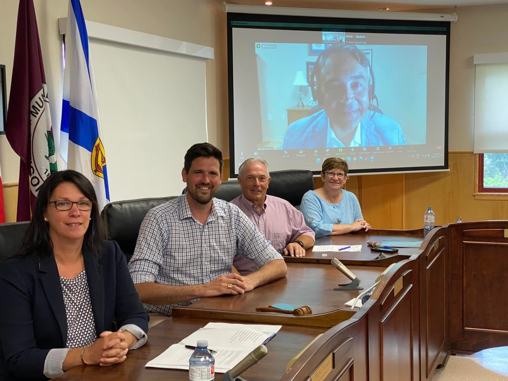 四个人坐在一张桌子前看着相机。背景是屏幕上的一个男人