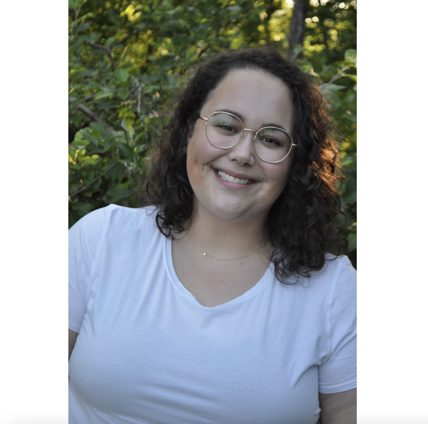 一个黑头发、戴着眼镜、穿着白色t恤的女人微笑着站在绿树和黄花前