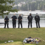 五名警察站在公园里，看着草地上一堆别人的东西，身后是水上的帆船