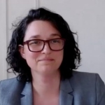 一位黑发及肩的女士戴着眼镜，穿着灰色西装外套。