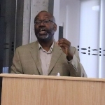 一个戴着眼镜，穿着米色夹克的黑人站在讲台上