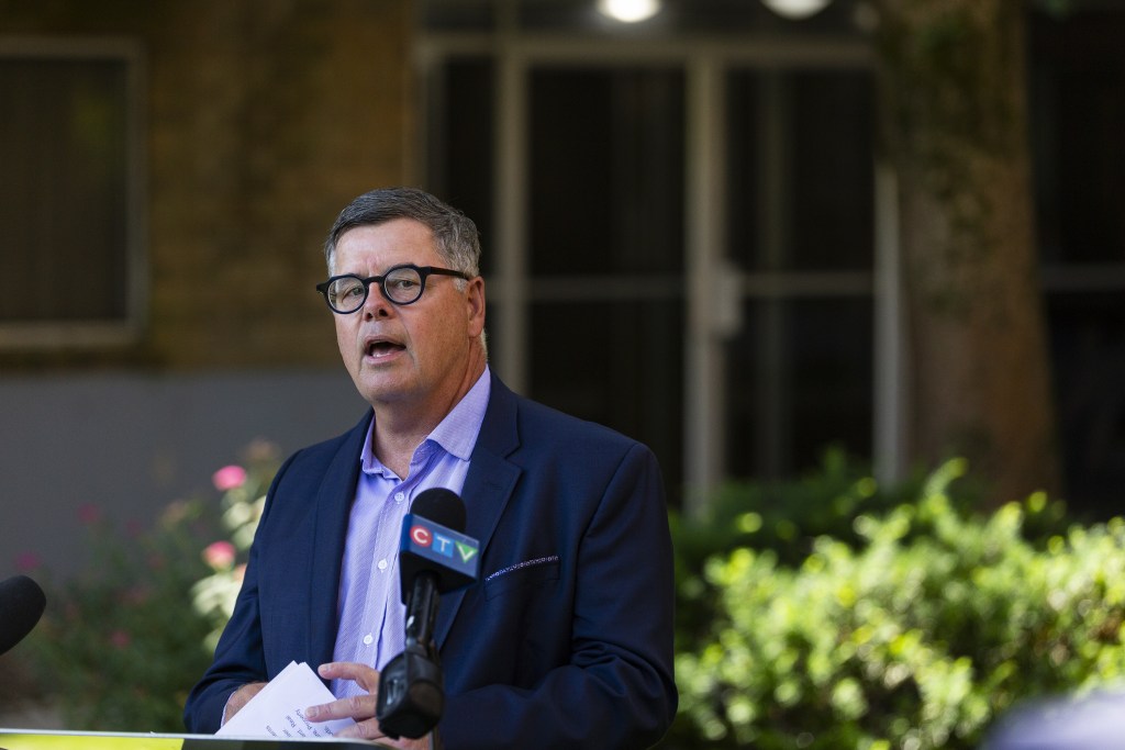 在一个阳光明媚的日子里，一名身穿西装、戴着厚边眼镜的男子在公寓大楼外的讲台上发言。