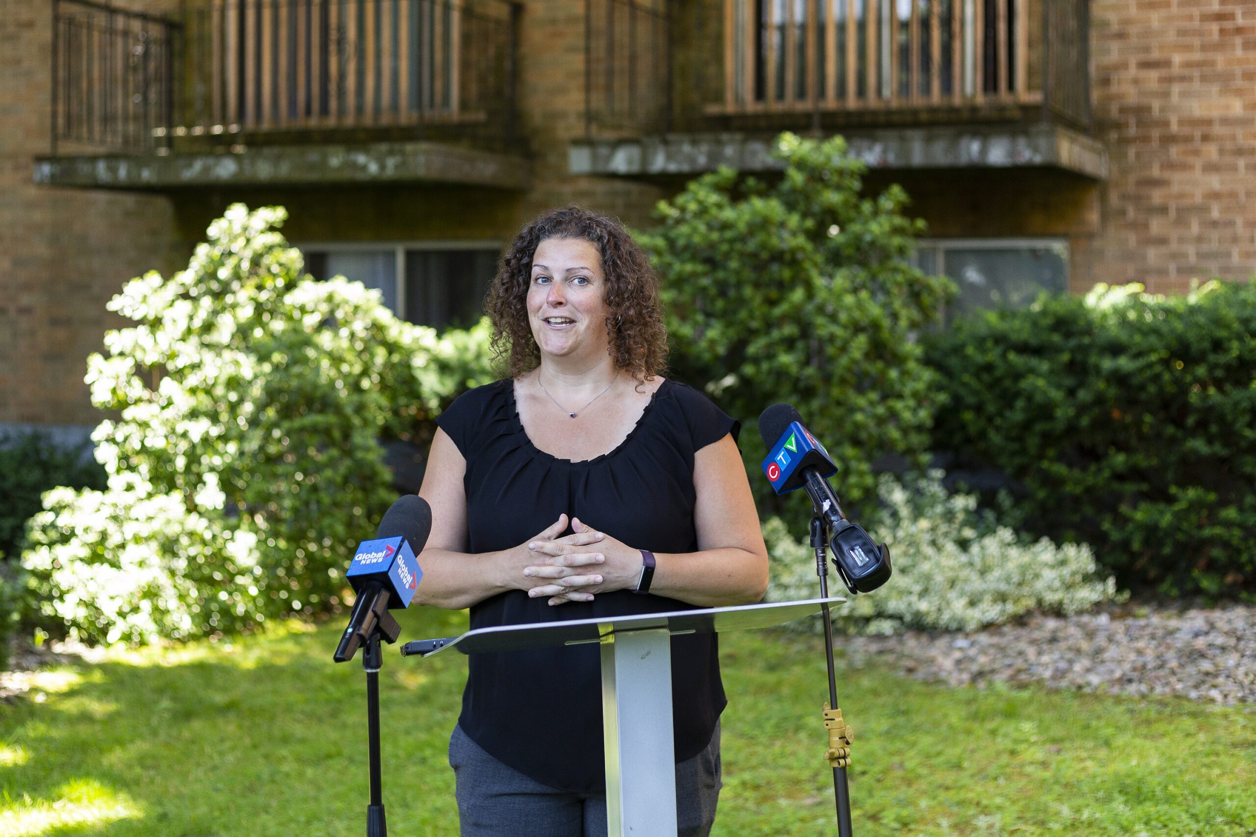 A woman speaks at a podium outside an apartment building on a sunny day.