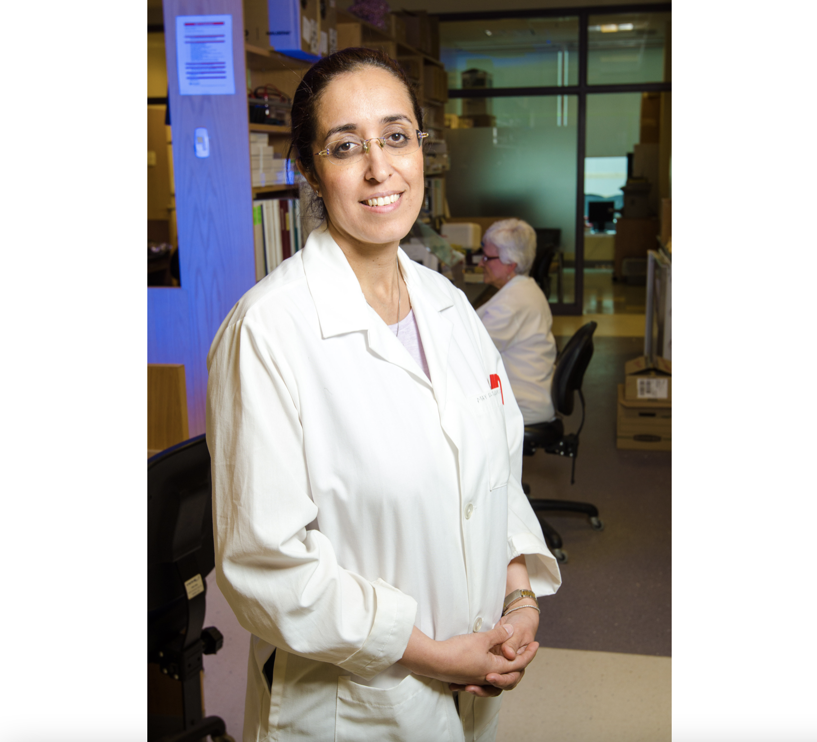 一个戴着眼镜、穿着白大褂、头发向后梳的女人微笑着站在实验室工作人员面前，对着镜头微笑。