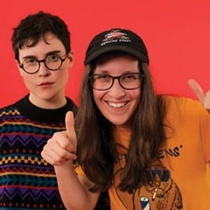 两个人站在一个橙色的屏幕前。左边的人留着黑色短发，戴着眼镜，穿着印花毛衣。右边的人留着棕色的长发，戴着一顶棒球帽，穿着芥末黄色的t恤。他们竖起了两个大拇指。