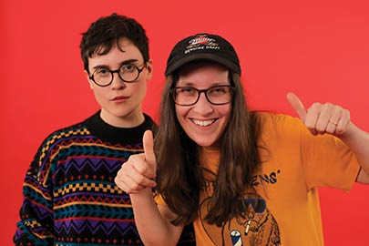 Two people stand in front of a bright orange background. The person on the left has short dark hair, glasses, and is wearing a printed sweater. The person on the right has long brown hair, is wearing a ball cap, and a mustard yellow t-shirt. They are giving two thumbs up.