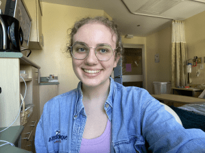 一个微笑的年轻女子戴着眼镜，在紫色衬衫外面穿了一件蓝色牛仔衬衫，看着以病房为背景的镜头。