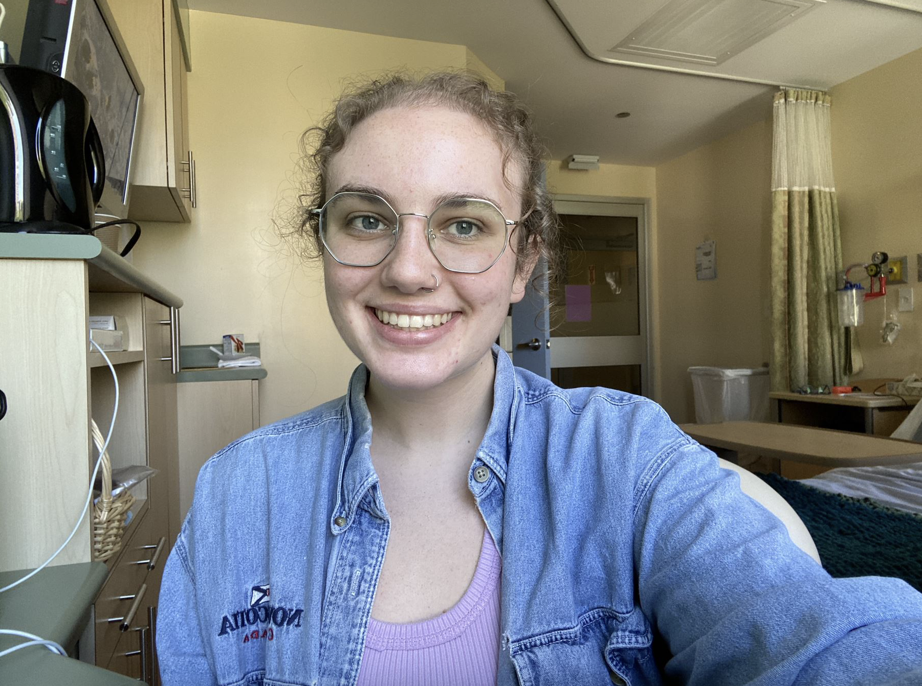 一名戴着眼镜、在紫色衬衫外面套着蓝色牛仔衬衫的年轻女子微笑着看着镜头，背景是医院的房间。