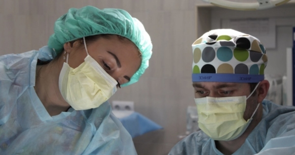 两名身穿蓝色外套、戴着黄色外科口罩的外科医生正在为一位看不见的病人做手术