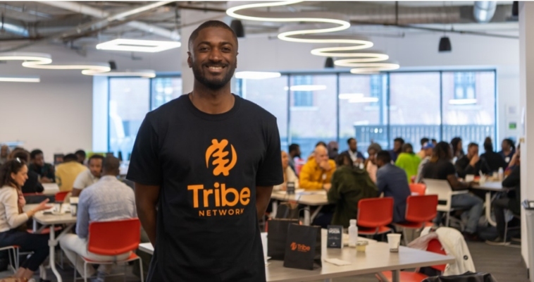 一个黑人站在会议室里，人们坐在桌旁。这个人穿着一件写着部落网络的t恤。