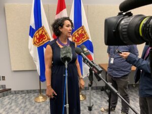 一个留着黑色短卷发、穿着黑色无袖连衣裙的女人站在几个麦克风前