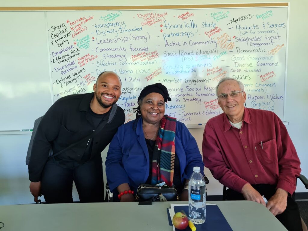 三个人坐在一张桌子前微笑着合影。在他们后面是一块写满了单词和短语的白板