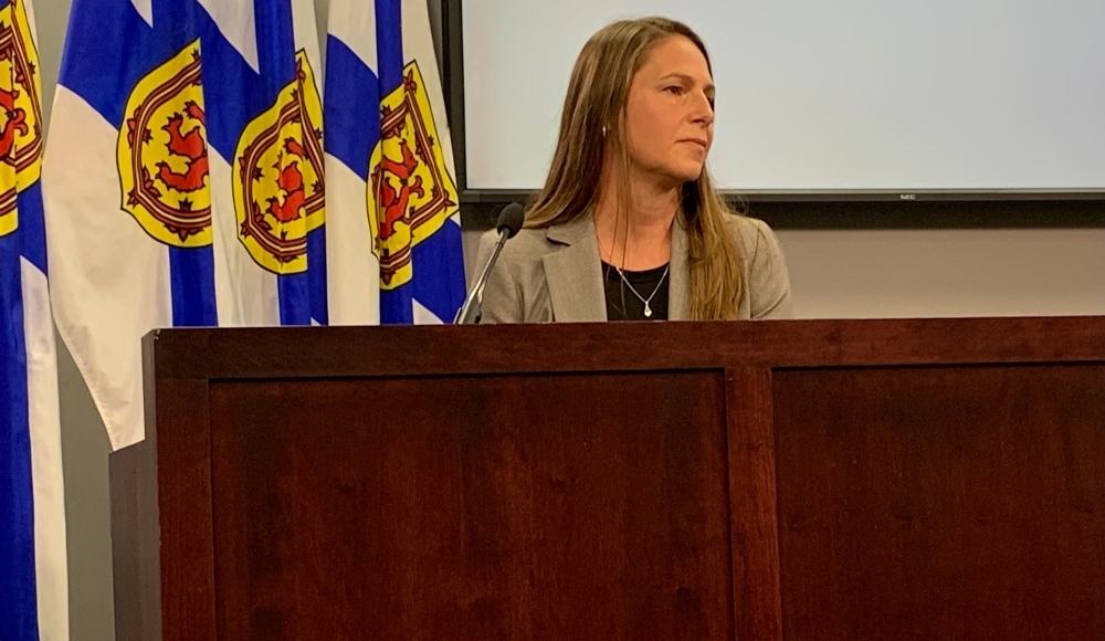 一名留着深金色长发、黑色上衣外穿米色西装外套的女子坐在一张深色木桌前，前面是一排新斯科舍省的旗帜