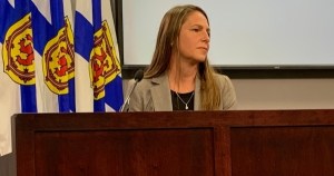 一名留着深金色长发、黑色上衣外穿米色西装外套的女子坐在一张深色木桌前，前面是一排新斯科舍省的旗帜