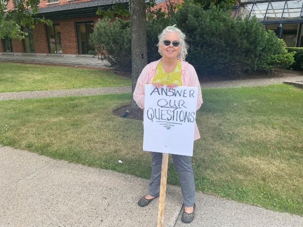 一位白发女子戴着墨镜，穿着粉色夹克和灰色裤子站在人行道上，手里举着一个牌子，上面写着“回答我们的问题”。