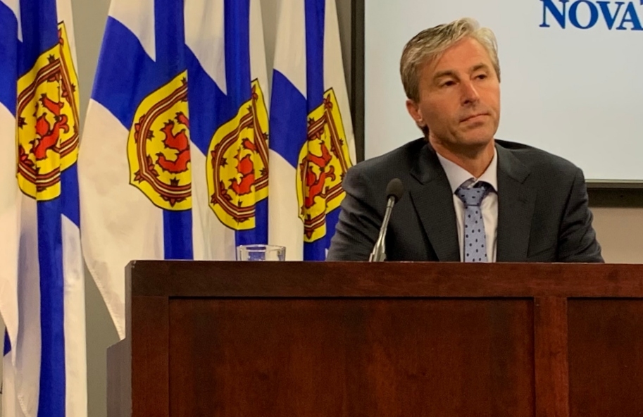 一名头发灰白、身穿深色西装、系着蓝色领带的男子坐在一张桌子前，前面是一排新斯科舍省国旗。