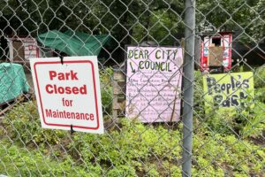 从一个写着“公园关闭维修”的栅栏里可以看到一个以前无家可归者的营地。栅栏后面还有其他标志，“亲爱的市议会”和“人民公园”。
