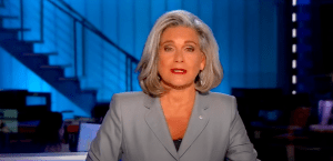 一位白发苍苍、身穿灰色西装的中年白人女性坐在蓝色背景的新闻台前