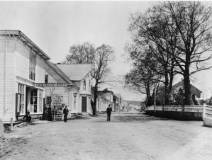 一个乡村的街景的黑白照片。一条肮脏的街道两旁矗立着几座建筑物，人们穿着维多利亚时代的服装在建筑物外散步或站立。