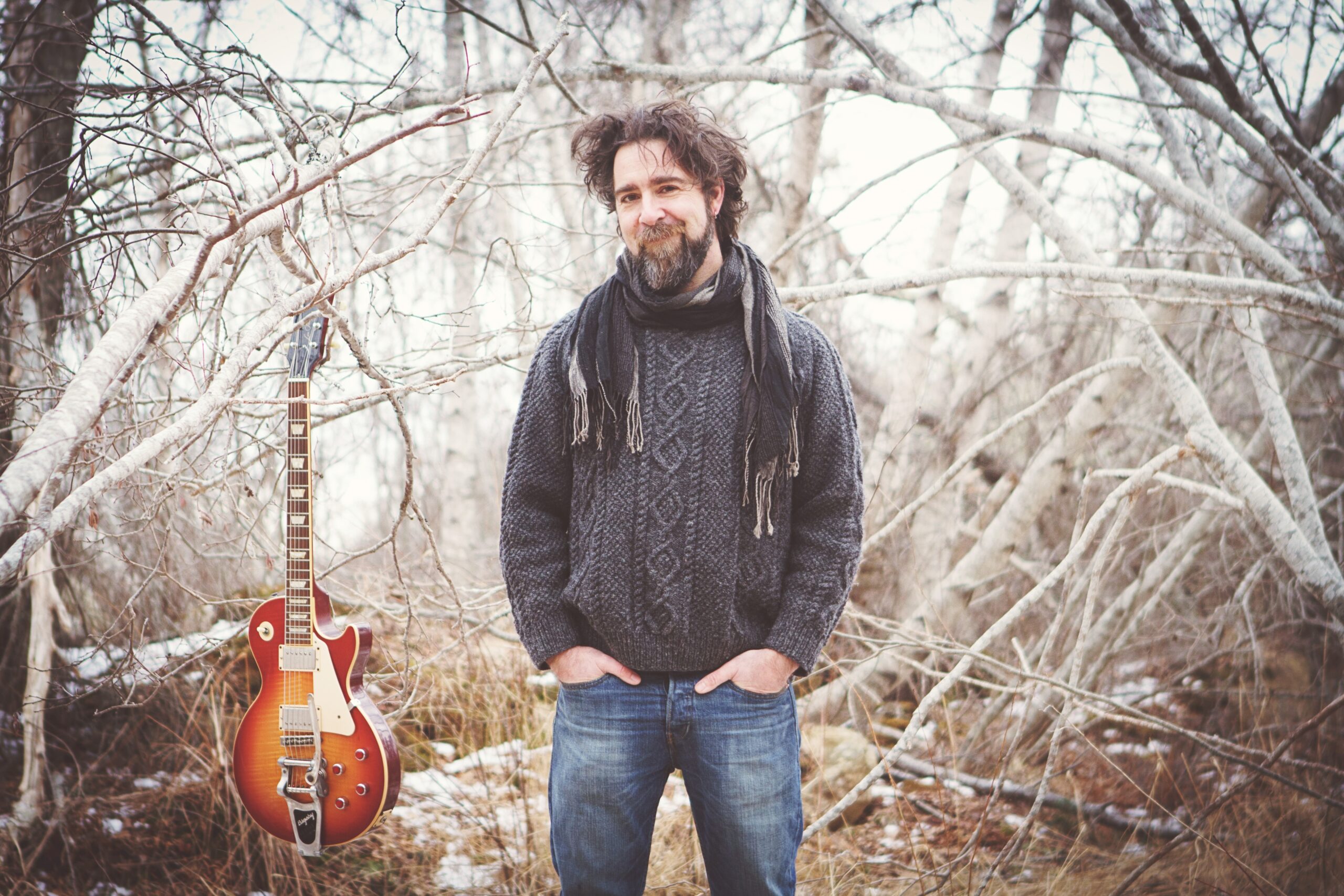 一个留着灰色斑点胡子的中年白人男子站在树林里。他穿着牛仔裤和毛衣。他身旁放着一把电吉他。