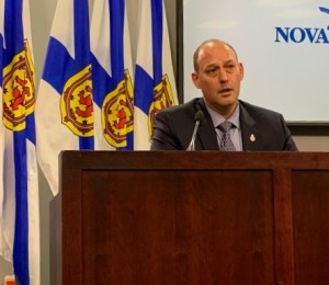 一个西装革履的秃头男人坐在桌子前。他身后是一排新斯科舍省的国旗