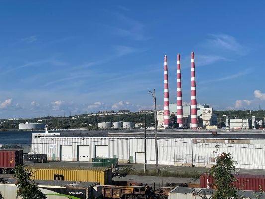 在蓝天和几朵云的背景下，可以看到三个红色和白色的堆栈。前景是一个船坞和轨道上的有轨电车。