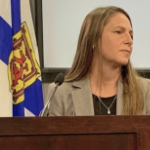 一位留着深金色长发的白人妇女在新斯科舍省国旗前的讲台上讲话