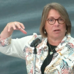 一位留着长及下巴的棕色头发、身穿印有彩虹斑马图案的衬衫的白人妇女在麦克风前做手势讲话