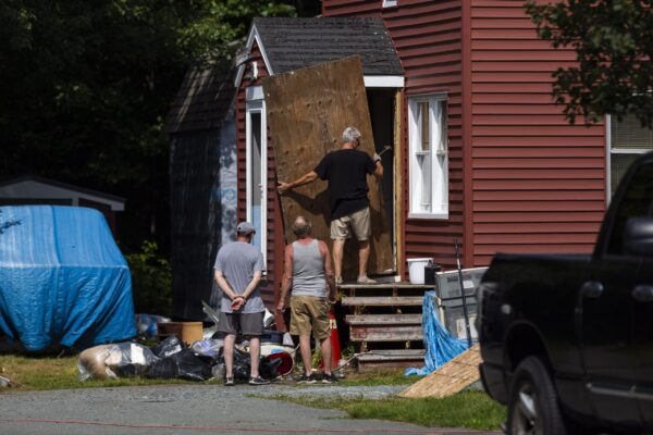 一个人拿着锤子，在两个人的注视下移动一块胶合板来盖住一扇门。房子是暗红色的，车道上停着一辆旧卡车，房子旁边还有一辆被蓝色防水布盖着的车和垃圾