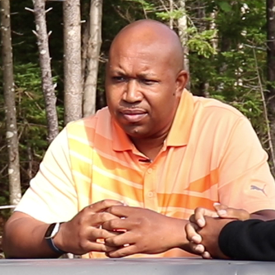 一个穿橙色衬衫的黑人坐在那里，双手交叉着手指