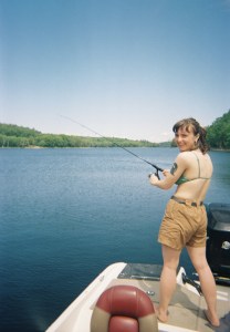 在一个阳光明媚的日子里，一个穿着比基尼上衣和短裤的女人在湖上的船上拿着钓竿。她背对着摄像机。她正转身对着镜头微笑