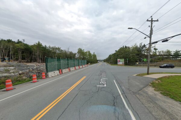 一张街景截图显示了一个阴天的十字路口。在左边，在混凝土球衣屏障上有绿色栅栏，底部有橙色的交通锥。