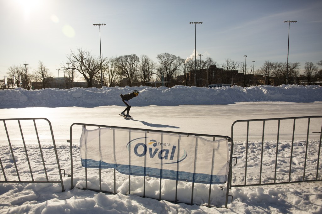 在一个阳光明媚的日子里，一名速滑运动员将手臂收在身后，在椭圆形场地上滑冰。背景是成堆的雪，前景的栅栏上有一个牌子:“Emera Oval”。