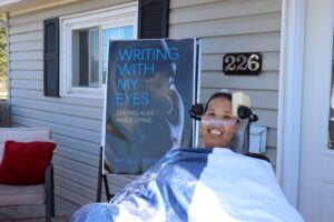 一个坐在轮椅上，裹着毯子微笑的黑人妇女。她的旁边是她的书的封面海报，用我的眼睛写作:在死亡中活着